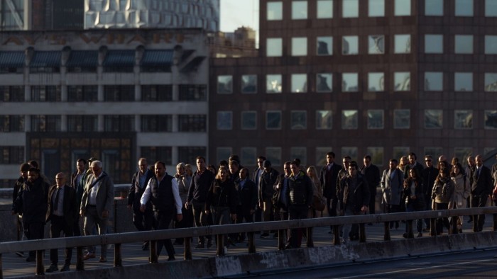 A large group of commuters walk across London Bridge in the early morning, with office buildings visible in the background.