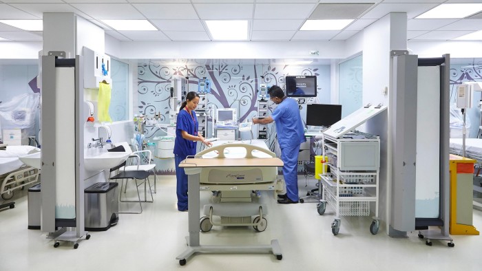 Two healthcare workers in blue scrubs prepare a hospital bed in a modern, well-equipped private ward.