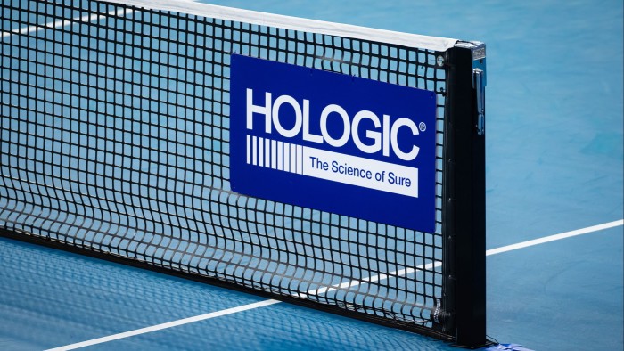 Hologic logo and slogan displayed on a blue sign attached to a tennis net on a hard court