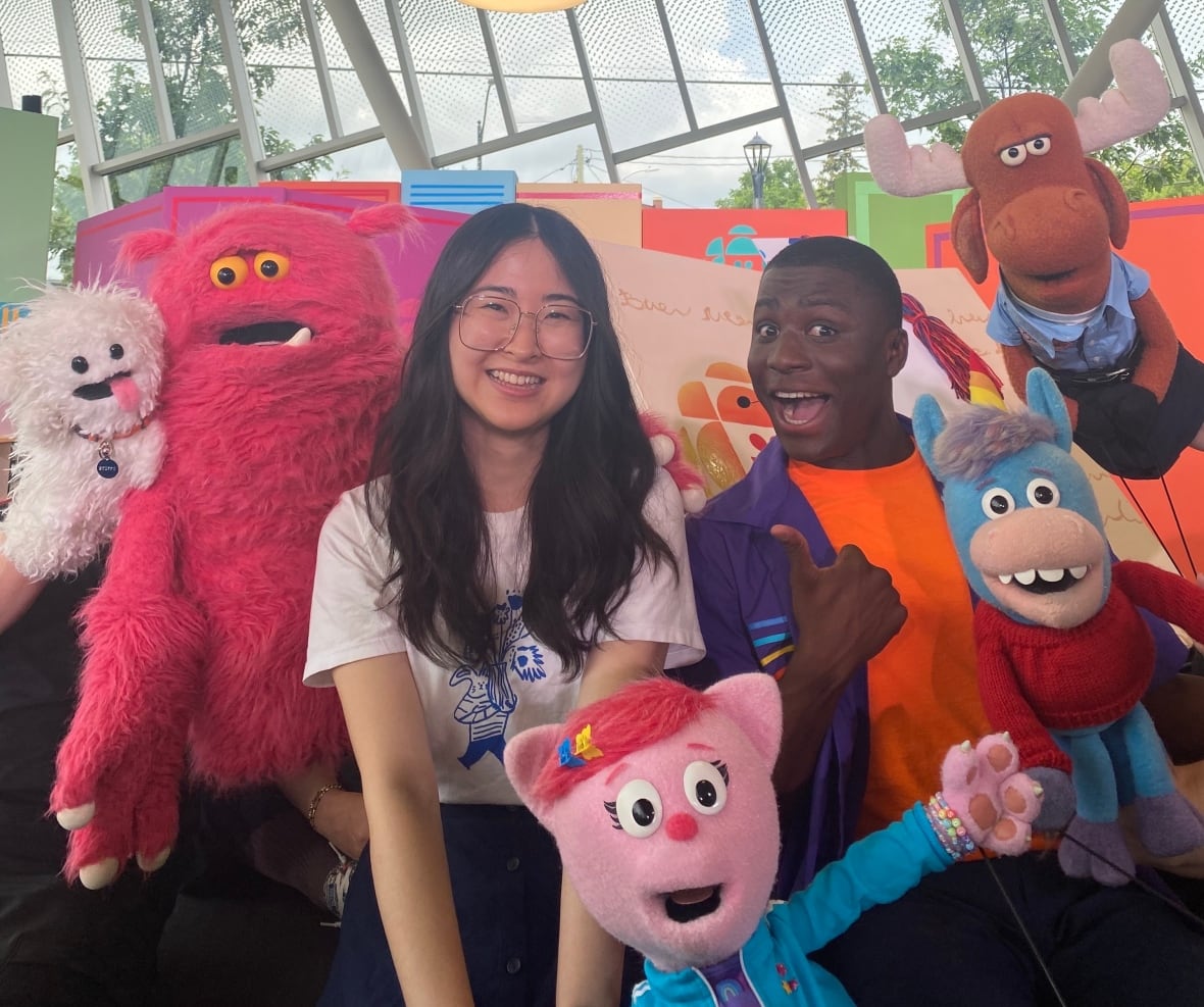 A woman and man smile at the camera, surrounded by CBC Kids puppets.