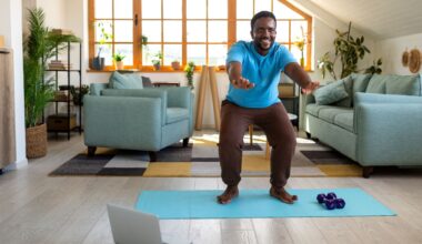 man performing a squat with arms outstretched in front on a blue exercise mat. there's a laptop on the floor in front of him and a living room setting with two sofas and a large window behind him