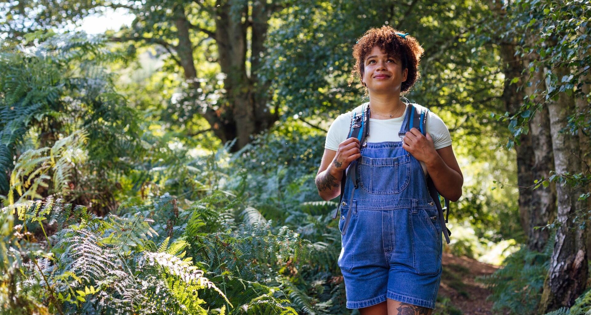 woman wearing denim short dungarees and a white tshirt walking in a forest setting.