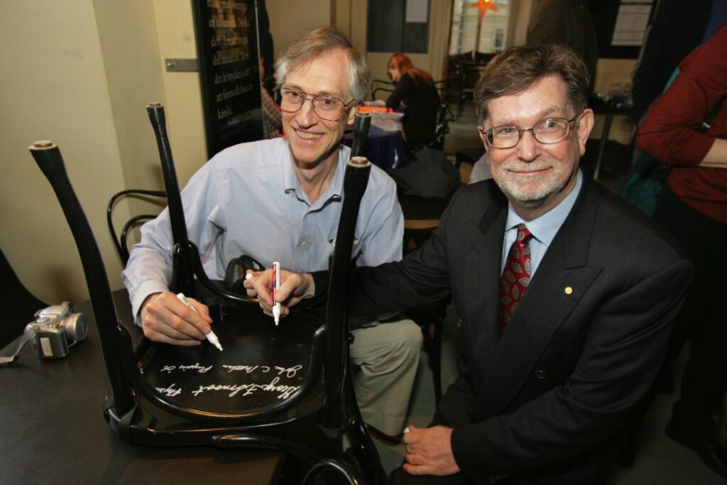 two men signing their names on the underside of a black chair