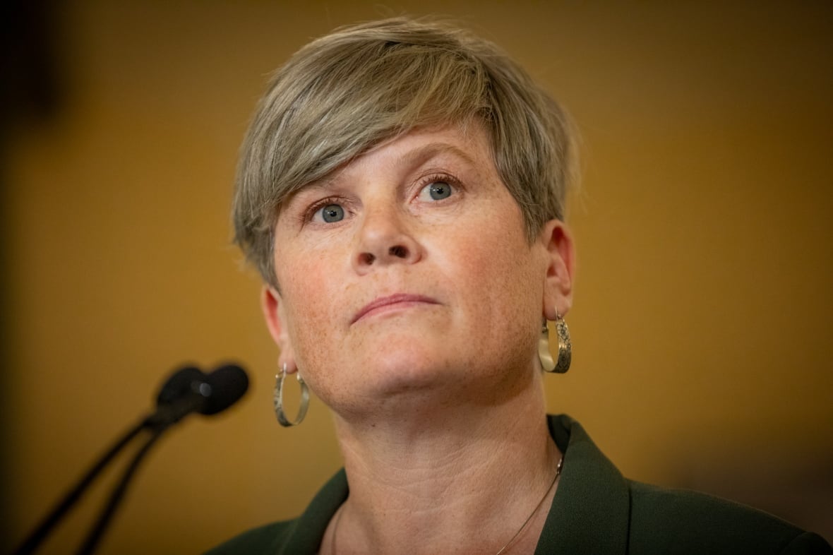 A white woman with short hair and hooped earrings is seen at a mic.