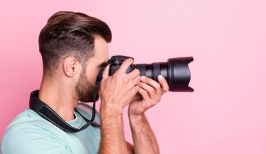 A man with short brown hair and a beard is holding a professional camera up to his face, taking a photo against a pink background. He is wearing a mint green t-shirt and has a camera strap around his neck.