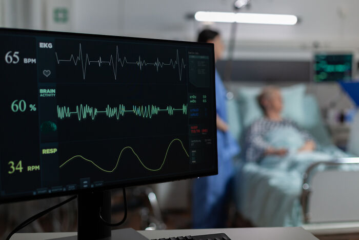 Hospital patient monitoring screen showing vital signs with nurse and elderly patient blurred in the background, highlighting terrifying symptoms.