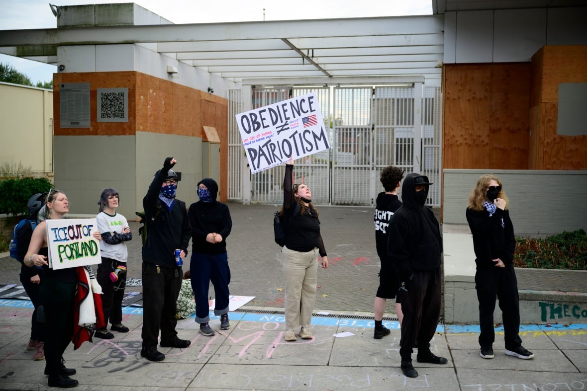 Protesters stand outside a gated building, with one person holding a sign saying 'Obedience [does not equal] Patriotism.'