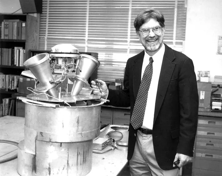 B&W photo of a man a suit and tie standing by a metal apparatus