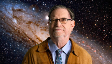 a man in a brown leather jacket, blue shirt and glasses, with the Andromeda Galaxy in the background.