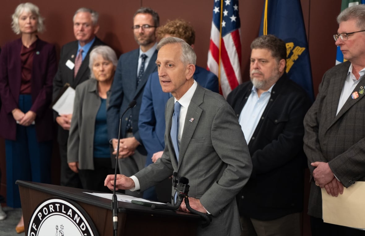 A group of men and women stand behind a grey-haired man, wearing a grey suit, who's speaking into a microphone.