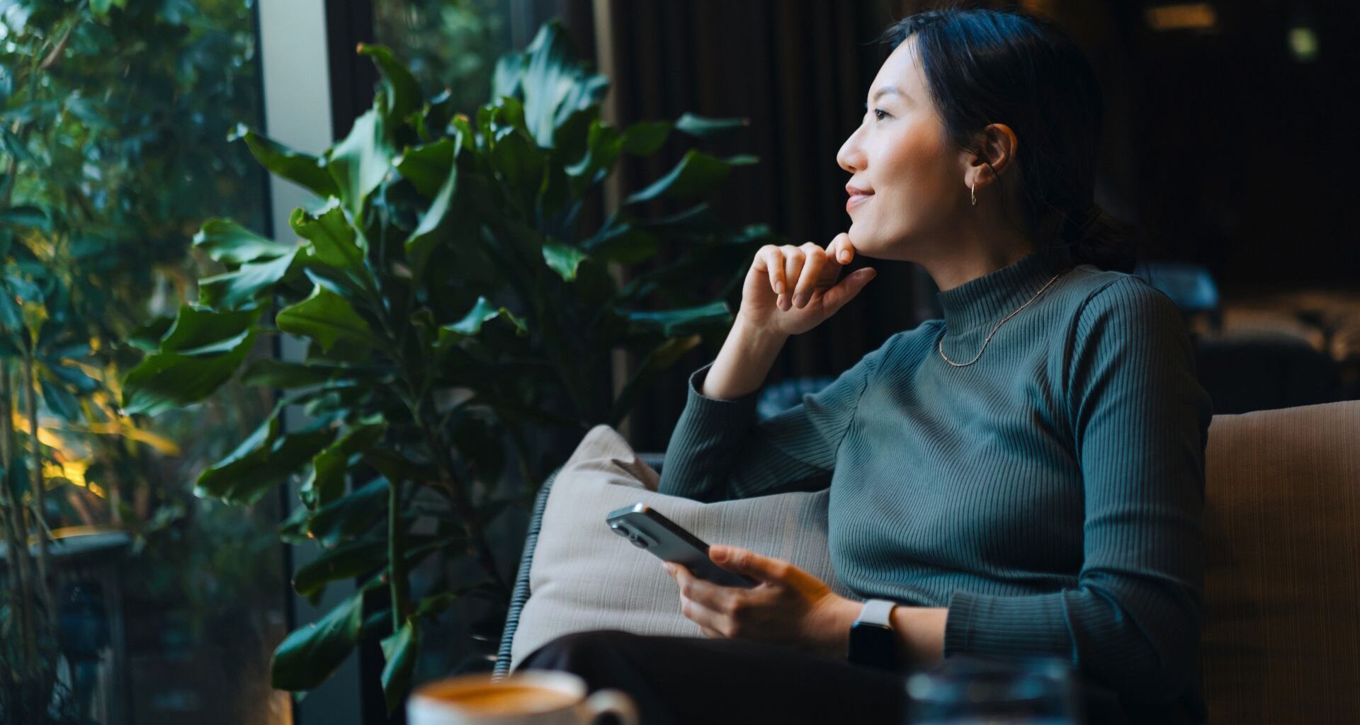 Woman smiling, holding phone and looking out of the window in a cafe, sitting on sofa after doing brain health exercises