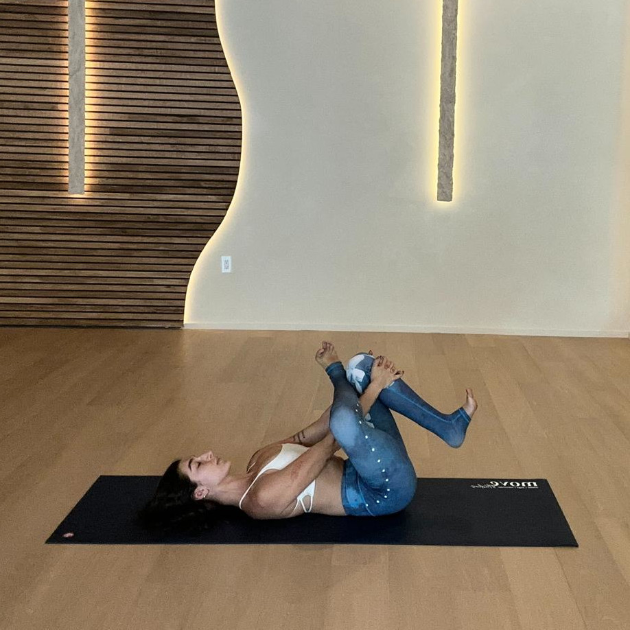 Yoga instructor Ilana Sabban from Move Studios performs a supine figure four stretch on yoga mat in a wood-floored studio. She is on her back, with her right foot crossed over her left knee, her hands looped around her left calf, pulling her legs towards her torso.