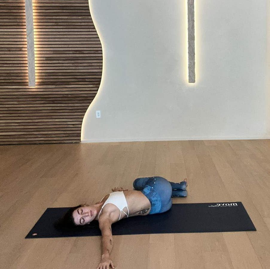 Yoga instructor Ilana Sabban from Move Studios performs a reclined spinal twist on a yoga mat in a wood-floored studio. She is lying with her arms out to the side, head facing the right, knees bent and legs together flopped over to the left side of her body.