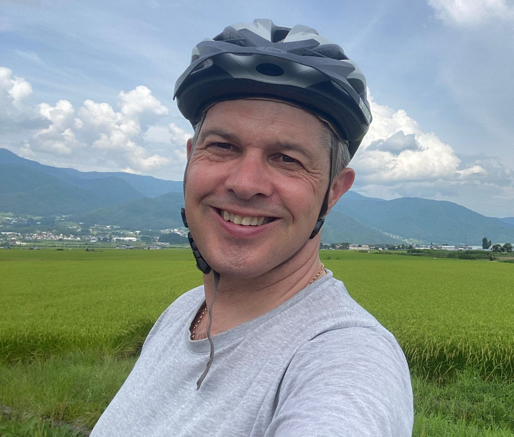 Neil Riley on holiday in Japan, wearing a cycling helmet and gray t-shirt, smiling in front of a green field, mountains and cloudy sky.