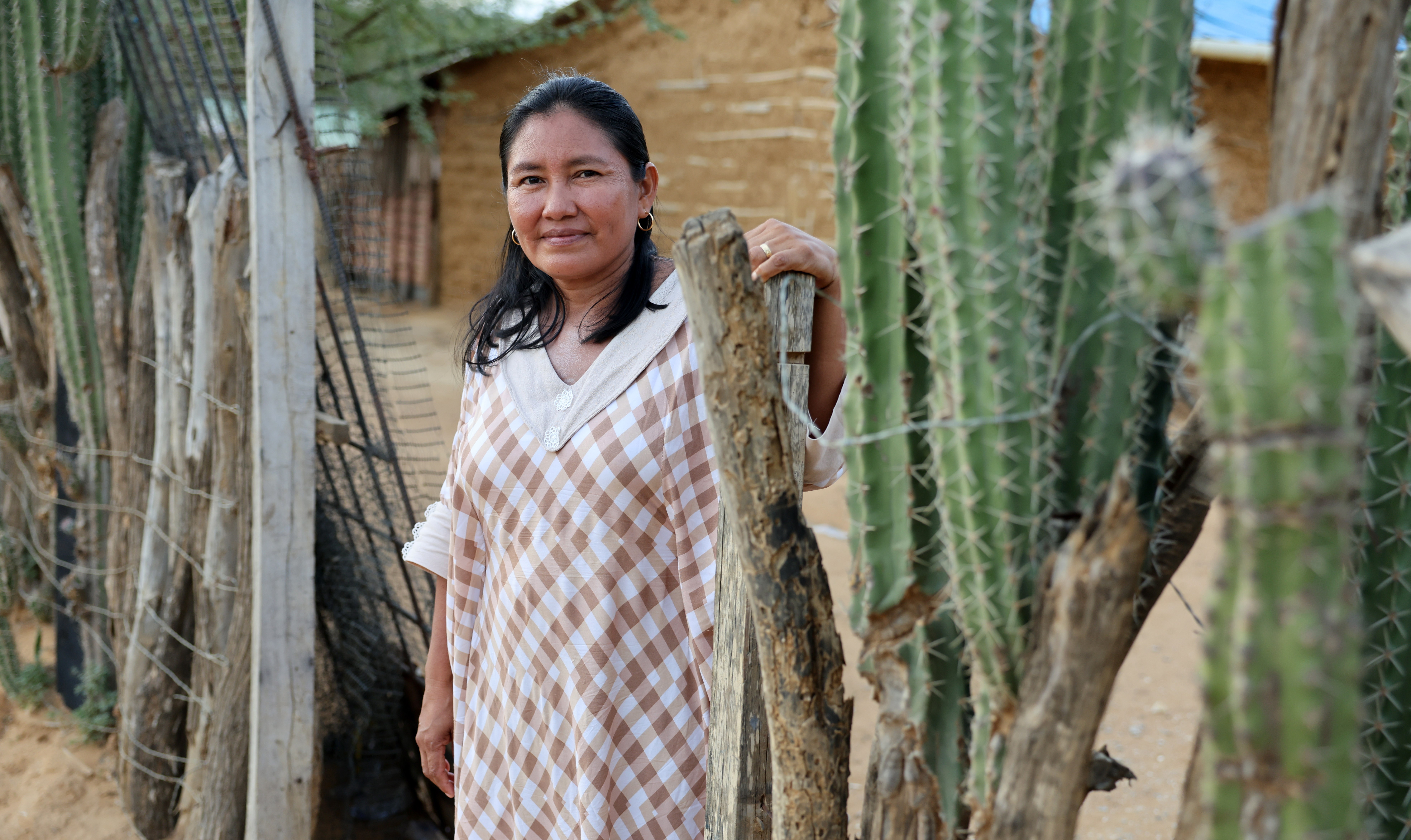 Rita Uriana, a Head Teacher and Community Leader, stands next to a wooden fence and large cacti, wearing a brown and white checkered dress.