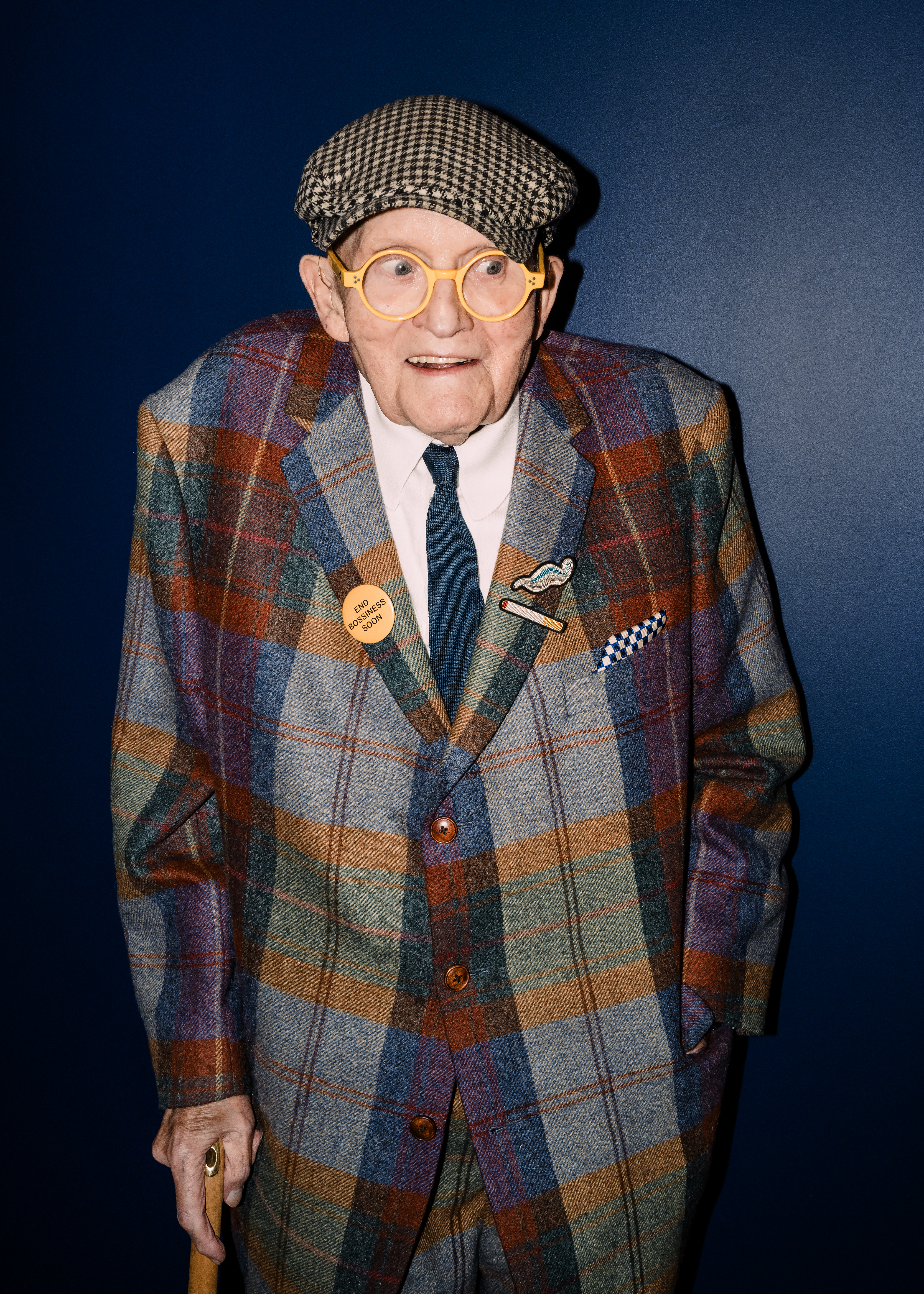 David Hockney at his latest exhibition, wearing a plaid blazer, a houndstooth cap, and yellow-framed glasses, holding a cane.