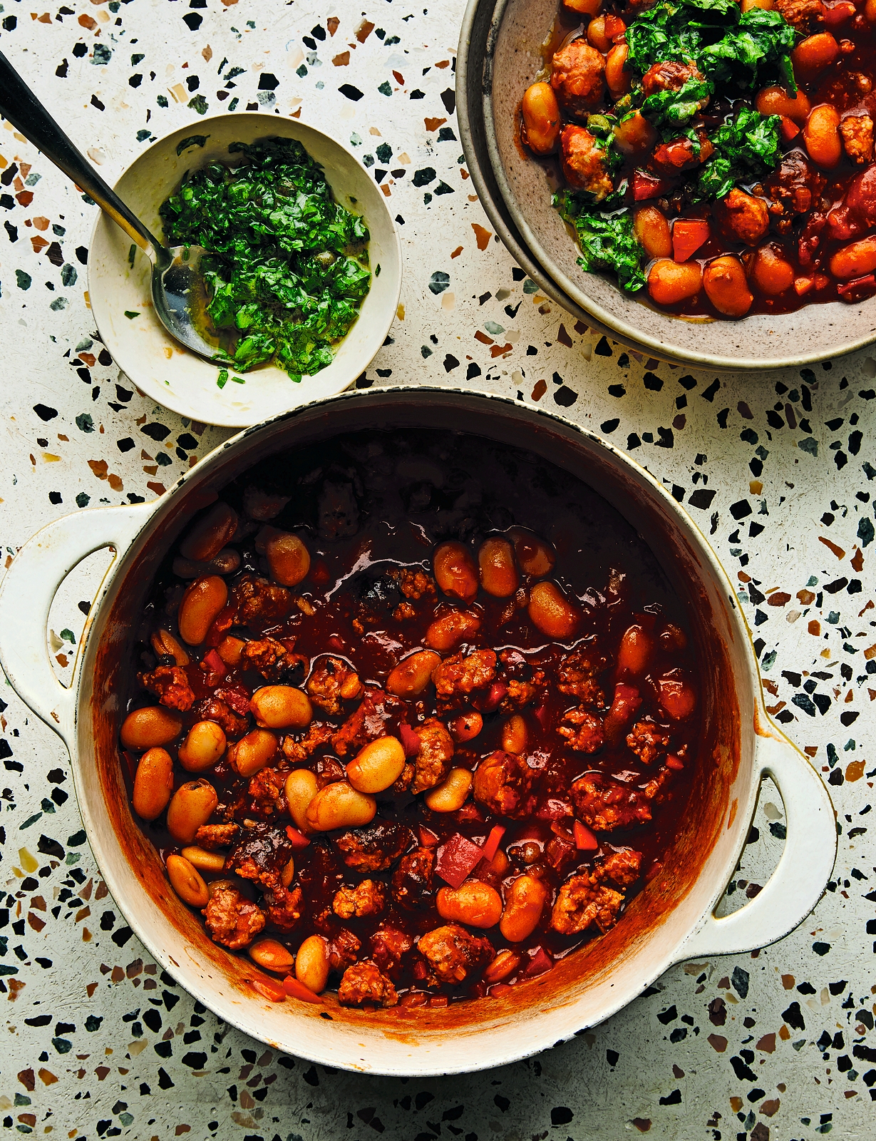 Sausage and butter beans in a pot and bowl, with a small dish of green herbs.