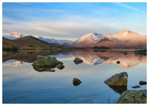 Frank McCafferty Snow-capped mountains rise under a soft blue sky, their peaks mirrored perfectly in a still, glass-like lake. Scattered rocks break the water’s surface, adding texture to the serene reflection.