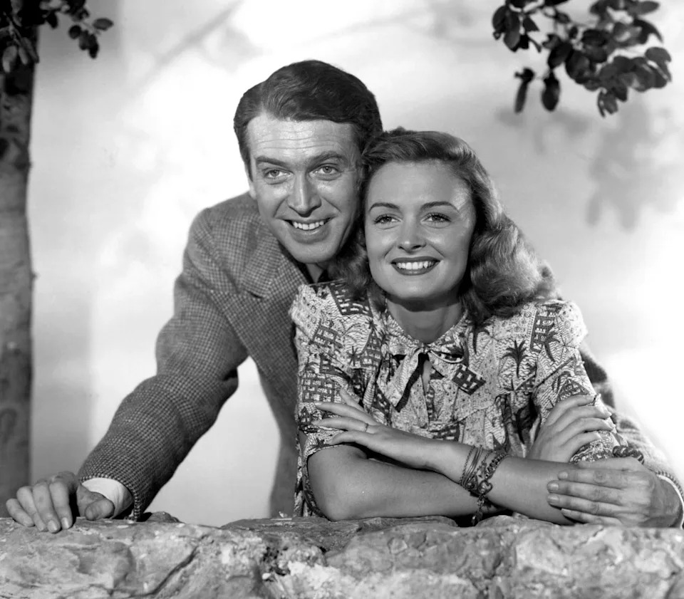 A classic black-and-white photo of a smiling man in a suit and a woman in a patterned dress leaning over a stone wall