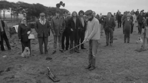 BBC A black-and-white still from the TV programme Nationwide in 1972. A crowd of people are watching a man in a flat cap with a stick attempt to hit a small ball-like object off a spring-based wooden trap.