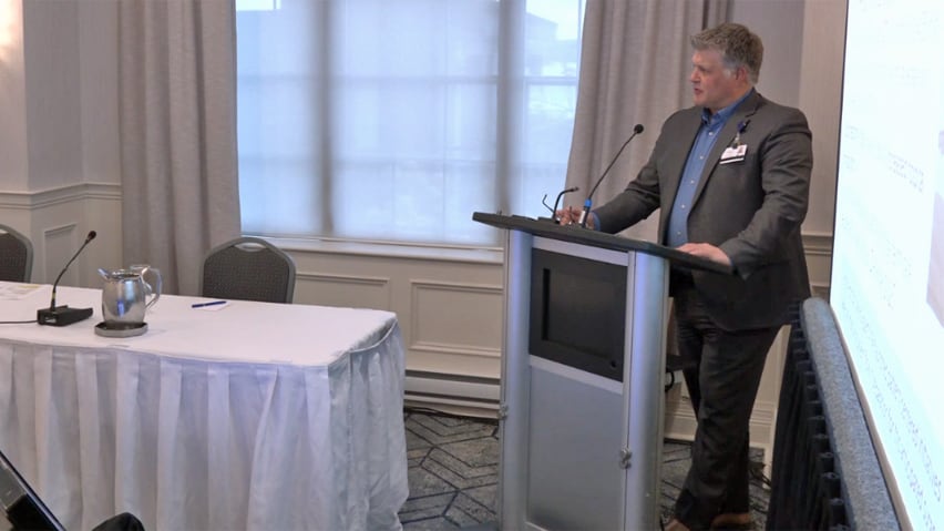 A man in a suit, standing at a podium, speaking into a microphone.