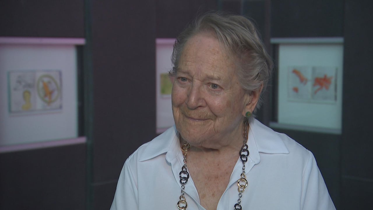 Older woman with short grey hair wearing white shirt. Behind her is a wall with sketches on display.