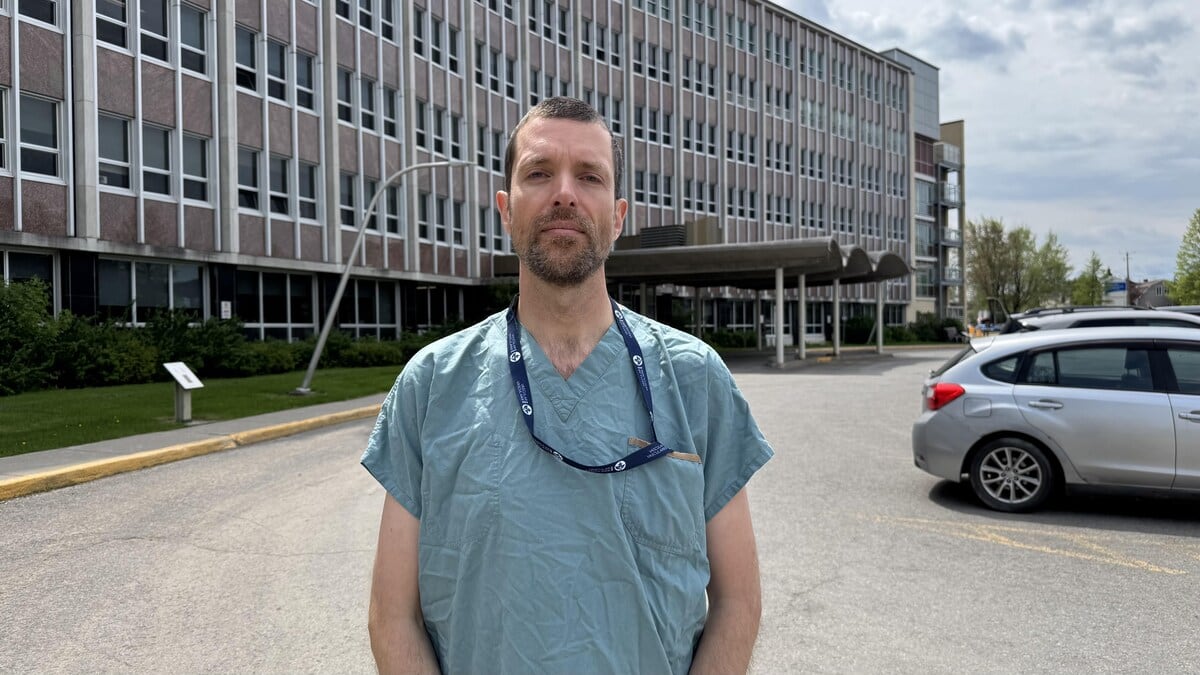 A white man wearing blue scrubs stands outside and looks into the camera.