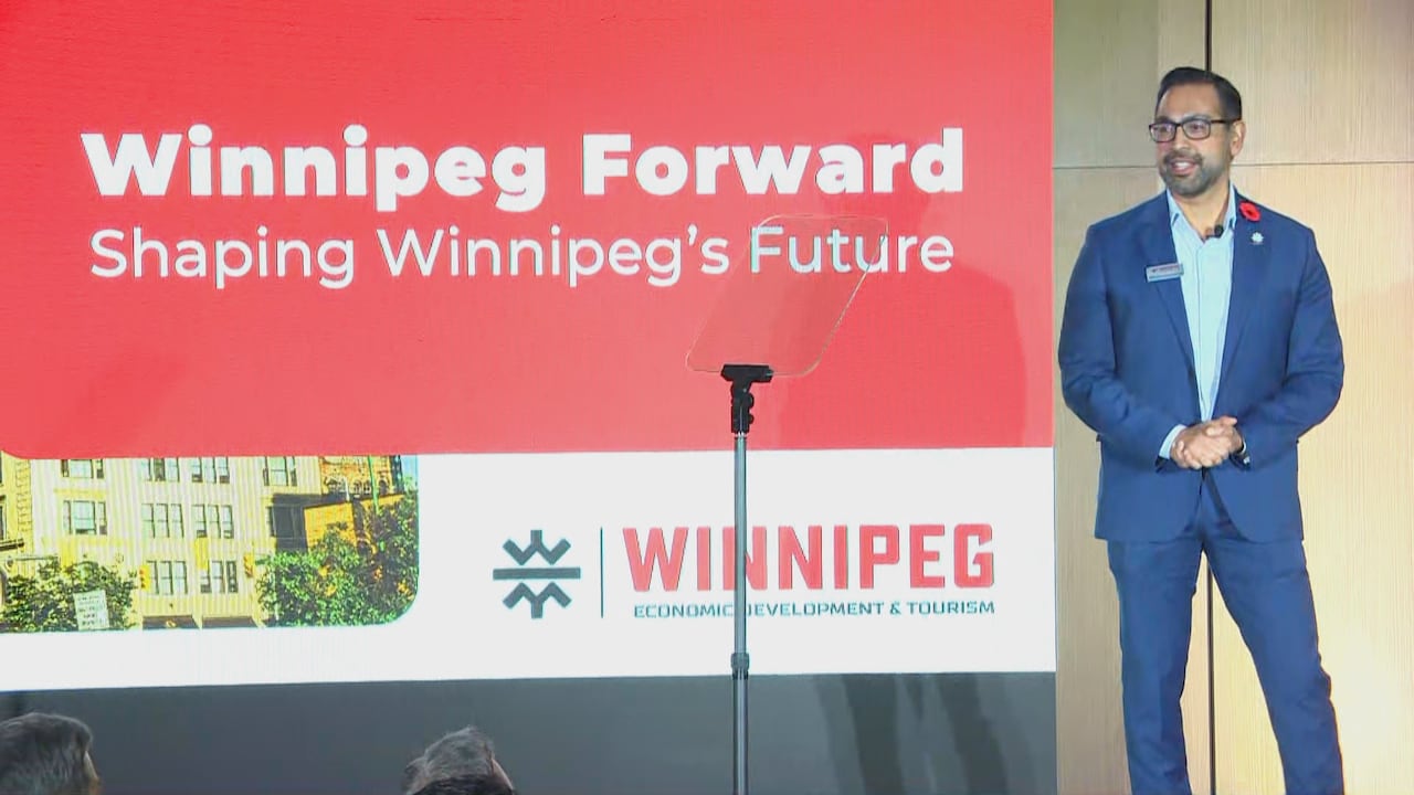 A man in a suit stands on a stage next to a large screen that says Winnipeg Forward.