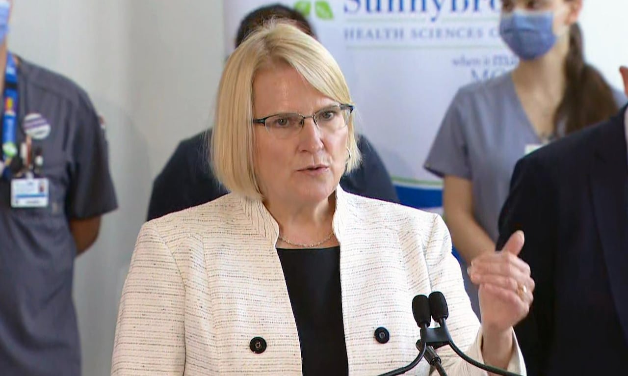 A woman with glasses stands at a podium and addresses media.