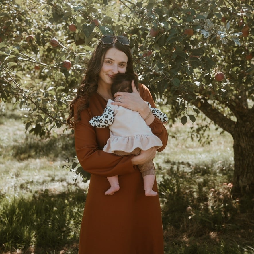 A woman holds an infant standing in front of an apple tree.