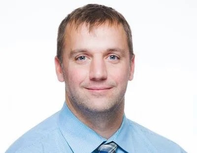 A man with short brown hair wearing a light blue dress shirt and a blue patterned tie, smiling at the camera against a plain white background.