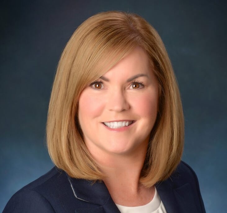 Headshot of a smiling woman with shoulder-length blonde hair, wearing a dark blue blazer over a white top, against a blue background