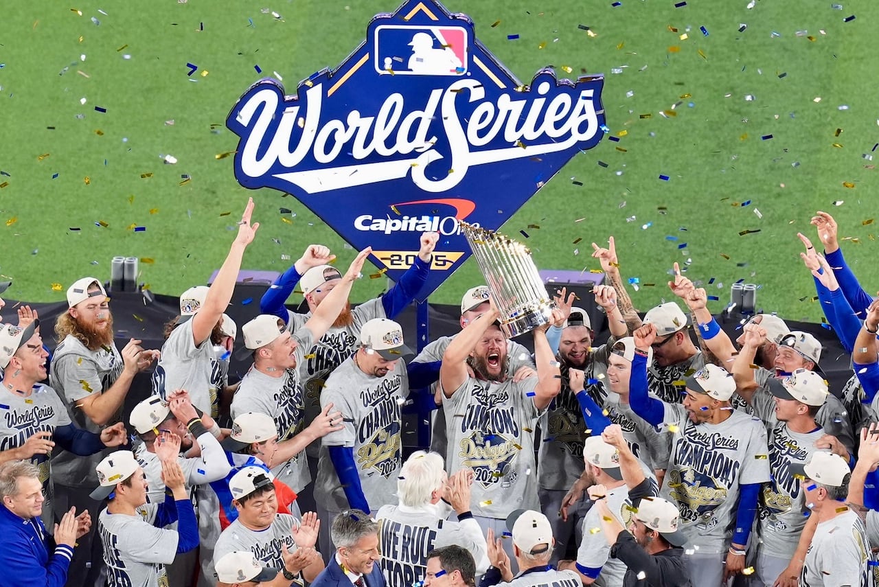 A professional baseball team celebrating with a trophy on a makeshift stage at a baseball field, with a sign behind them that reads: World Series.