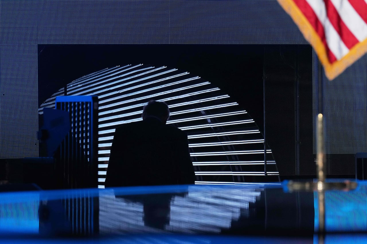 An elderly man walk away from a stage, his figure reflected partially on the stage floor.