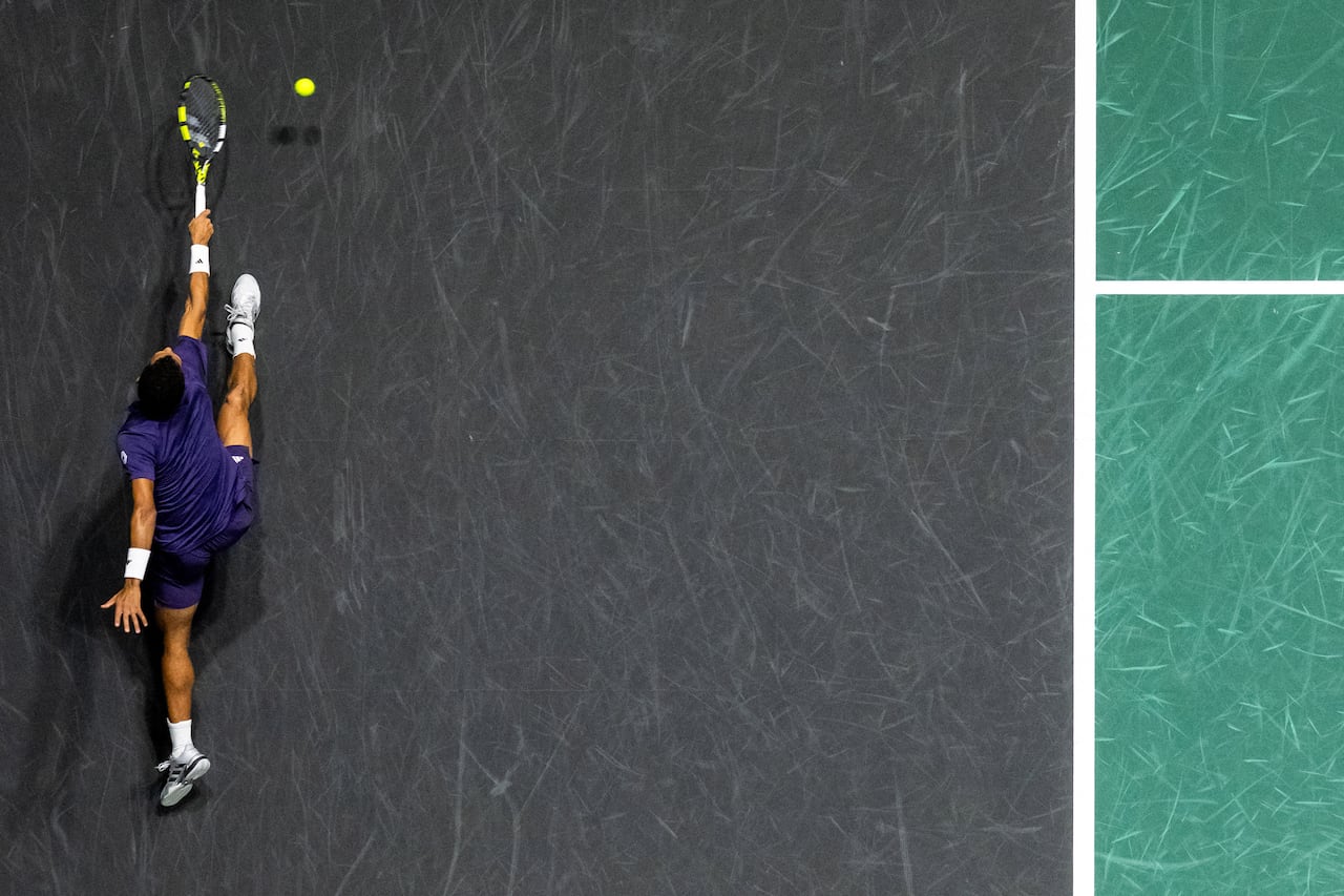 A bird's-eye view of a tennis player reaching to hit a ball with a backhand swing on a tennis court.