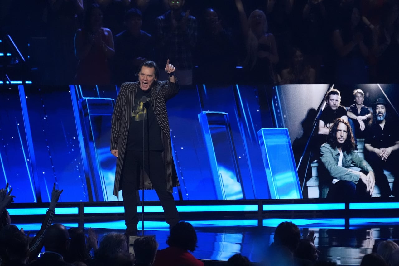 Jim Carrey stands on a blue-lit stage pointing out excitedly while he speaks to the audience