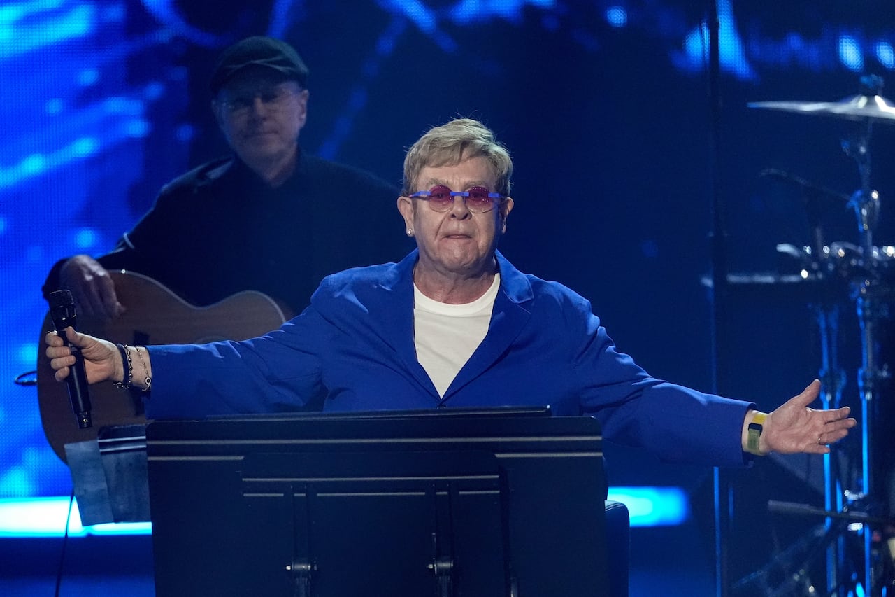 elton john licks his lips while holding his hands out wide while standing on stage