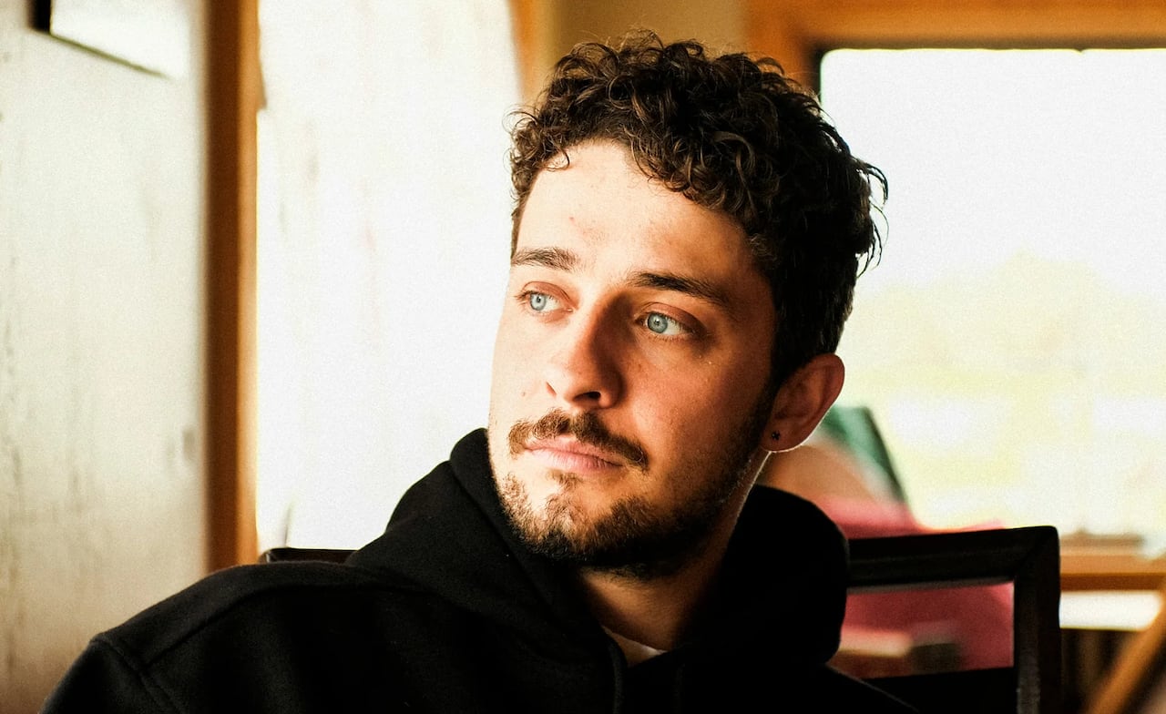 A young man with curly brown hair and blue eyes, wearing a black hoodie, looking off to the side in a warmly lit, casual indoor setting