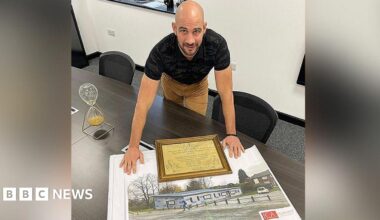 Dale Gaucas, wearing a black top and beige trousers, is leaning over a large photograph of the boxing club built in his old Darnhill estate.