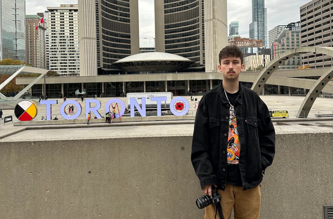 A man holding a camera standing in front of a Toronto sign and two tall, cement buildings