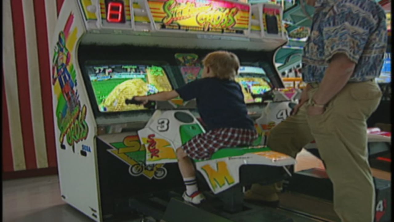 A young kid, with a man standing behind him, sitting on a motor bike arcade game.