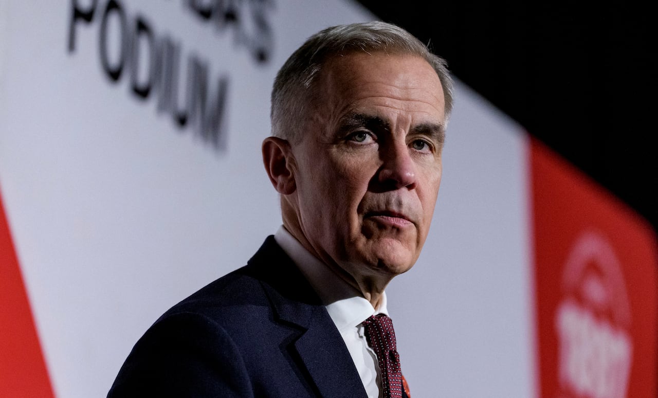 A white man speaks in front of a red and white screen.