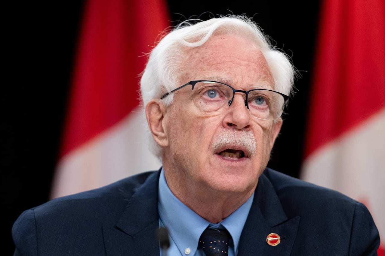 A man in a suit, wearing glasses, speaks to someone off screen. Canadian flags are seen in the background.