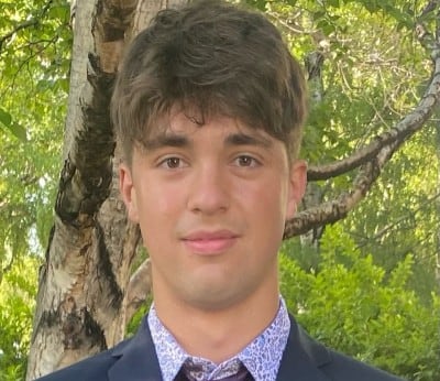  Young man stands in front of tree wearing patterned shirt, purple tie and navy blazer