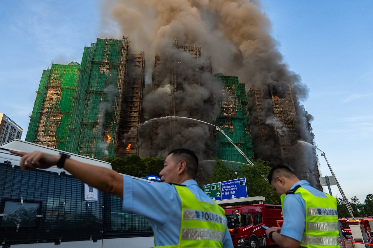 Two Asian men in reflective vests are shown in the foreground, with smoke pouring out of two buildings in the background that appear to be residential.