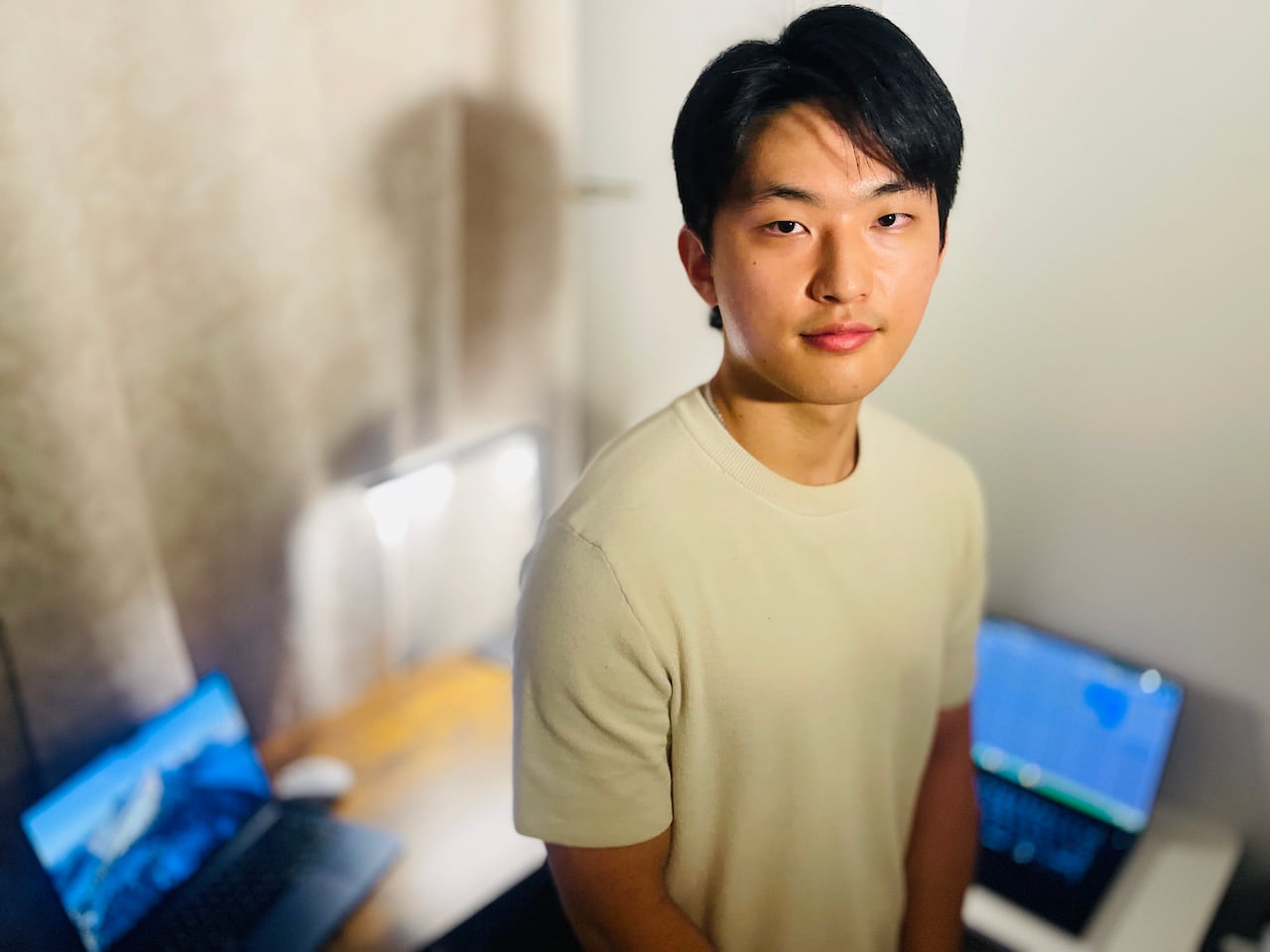 A young man in a beige tee-shirt stands in an indoor room, a desk and two computers seen out of focus behind him.