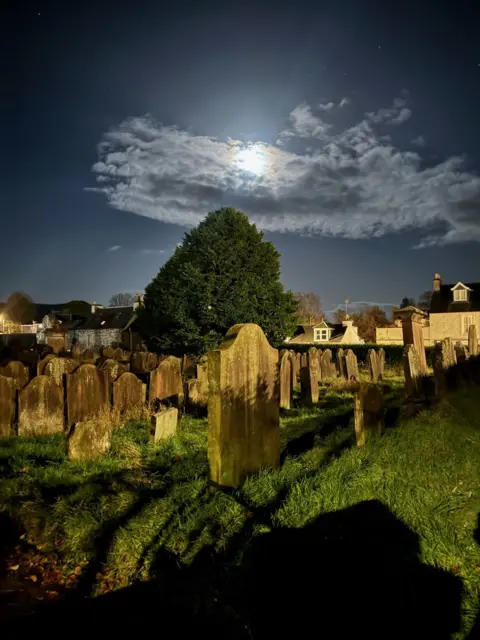 Fi Armstrong A moonlit graveyard is bathed in an eerie glow, with weathered headstones casting long shadows on the grass. A large tree stands prominently in the centre, framed by a cloud partially veiling the bright full moon. In the background, rooftops of houses peek through.