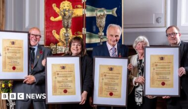 Recipients of the Freedom of the Borough stand from left - Cecil Page of the Kingfisher Boxing Club, Jayne Biggs of Norfolk Heart2Heart, Hugh Sturzaker of the Great Yarmouth Civic Society and members of the Great Yarmouth branch of The Samaritans. They are holding their scrolls of honour, mounted in frames, and stand before the staff and sceptre and the borough crest of Great Yarmouth. It is a shield painted red and blue divided vertically, with lion heads over the red segment, and herring fish tails over the blue, the heraldic image granted by King John in 1208.