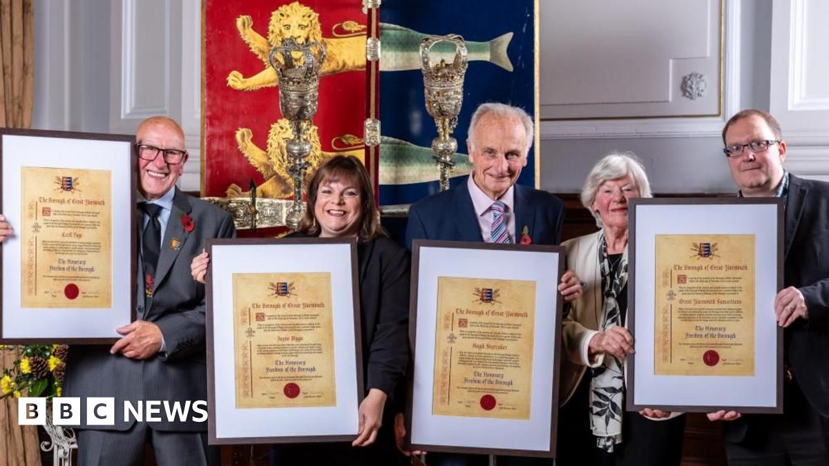 Recipients of the Freedom of the Borough stand from left - Cecil Page of the Kingfisher Boxing Club, Jayne Biggs of Norfolk Heart2Heart, Hugh Sturzaker of the Great Yarmouth Civic Society and members of the Great Yarmouth branch of The Samaritans. They are holding their scrolls of honour, mounted in frames, and stand before the staff and sceptre and the borough crest of Great Yarmouth. It is a shield painted red and blue divided vertically, with lion heads over the red segment, and herring fish tails over the blue, the heraldic image granted by King John in 1208.