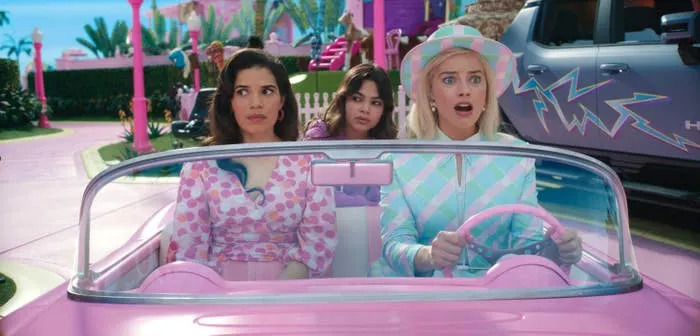 A scene from Barbie with America Ferrera, Ariana Greenblatt, and Margot Robbie in a convertible, one in a checkered outfit with a hat, and the others in patterned tops, look surprised while driving through a vibrant setting